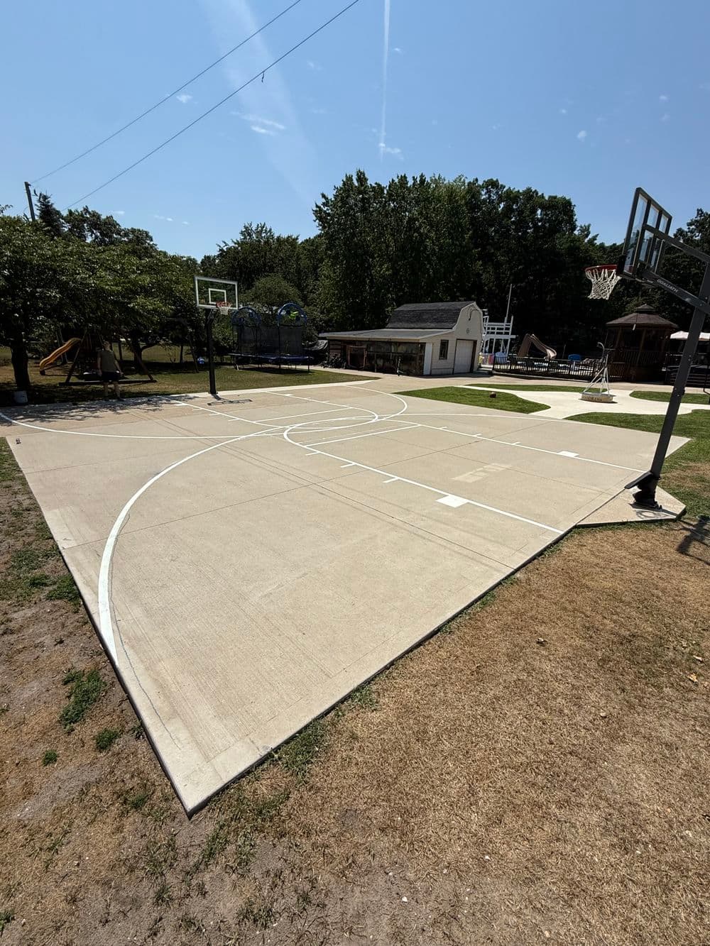 Outdoor basketball court with hoop, playground equipment, and green trees in sunny park setting.