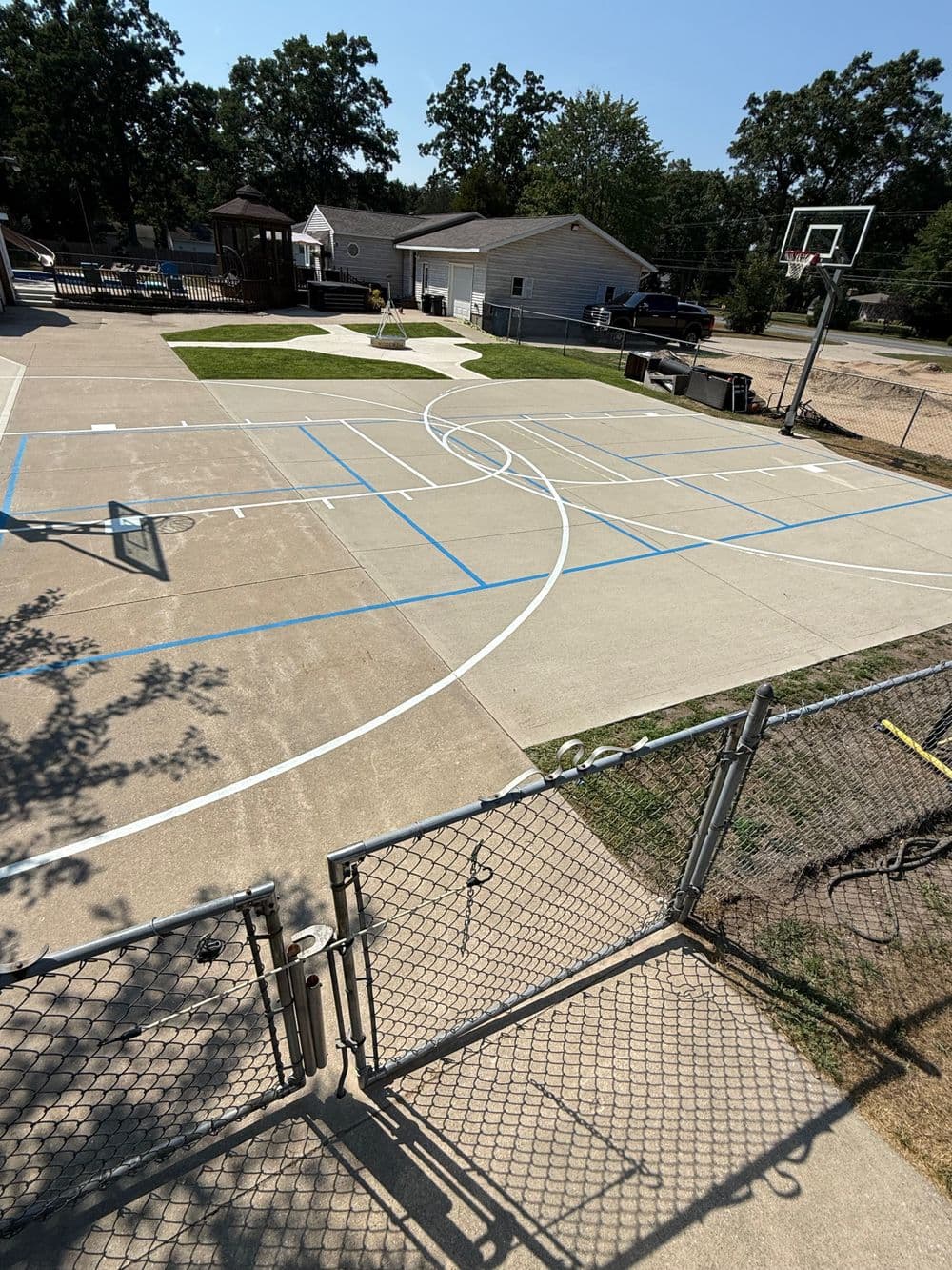 Basketball court with lines, fenced area, playground, and nearby houses on a sunny day.